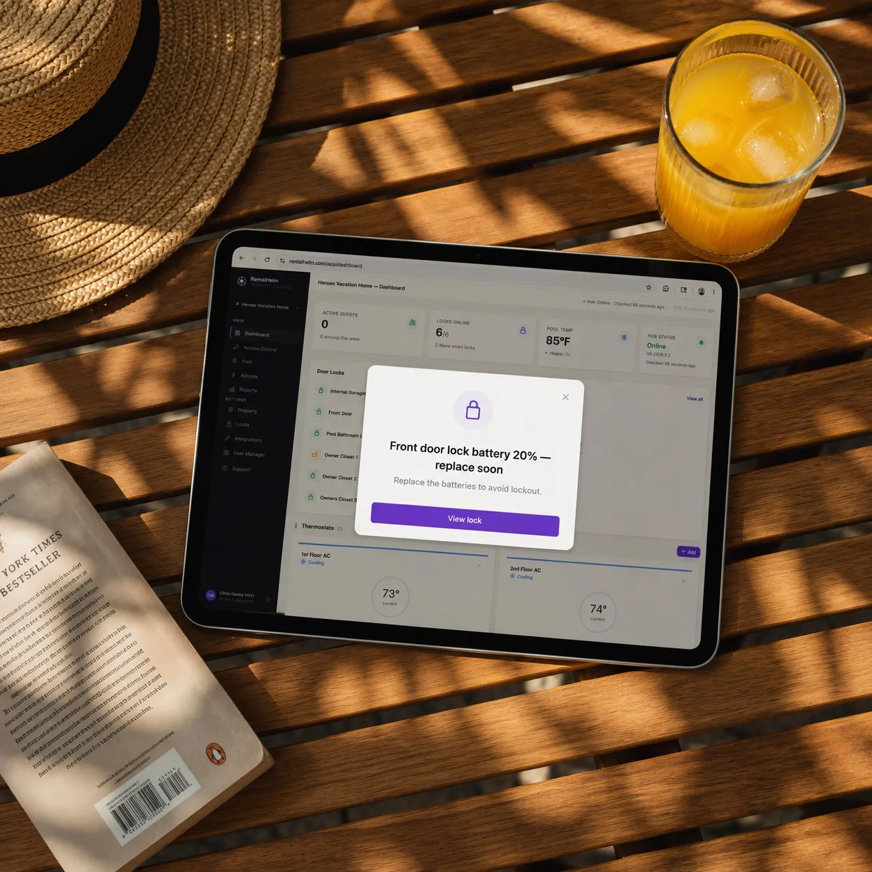 An iPad on a sunny patio table showing a RentalHelm 'Front door lock battery 20% — replace soon' alert, beside an orange juice and a straw hat